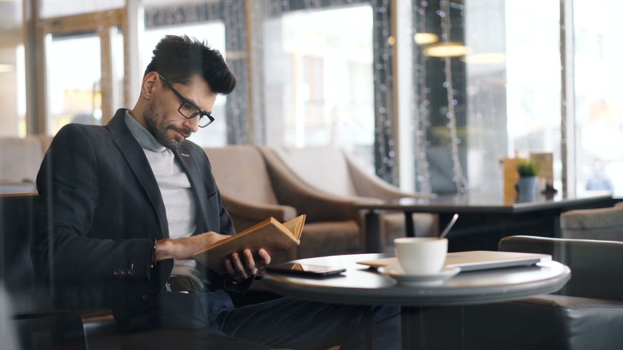 Focused businessman reading a book in a cozy, modern café setting with coffee nearby.